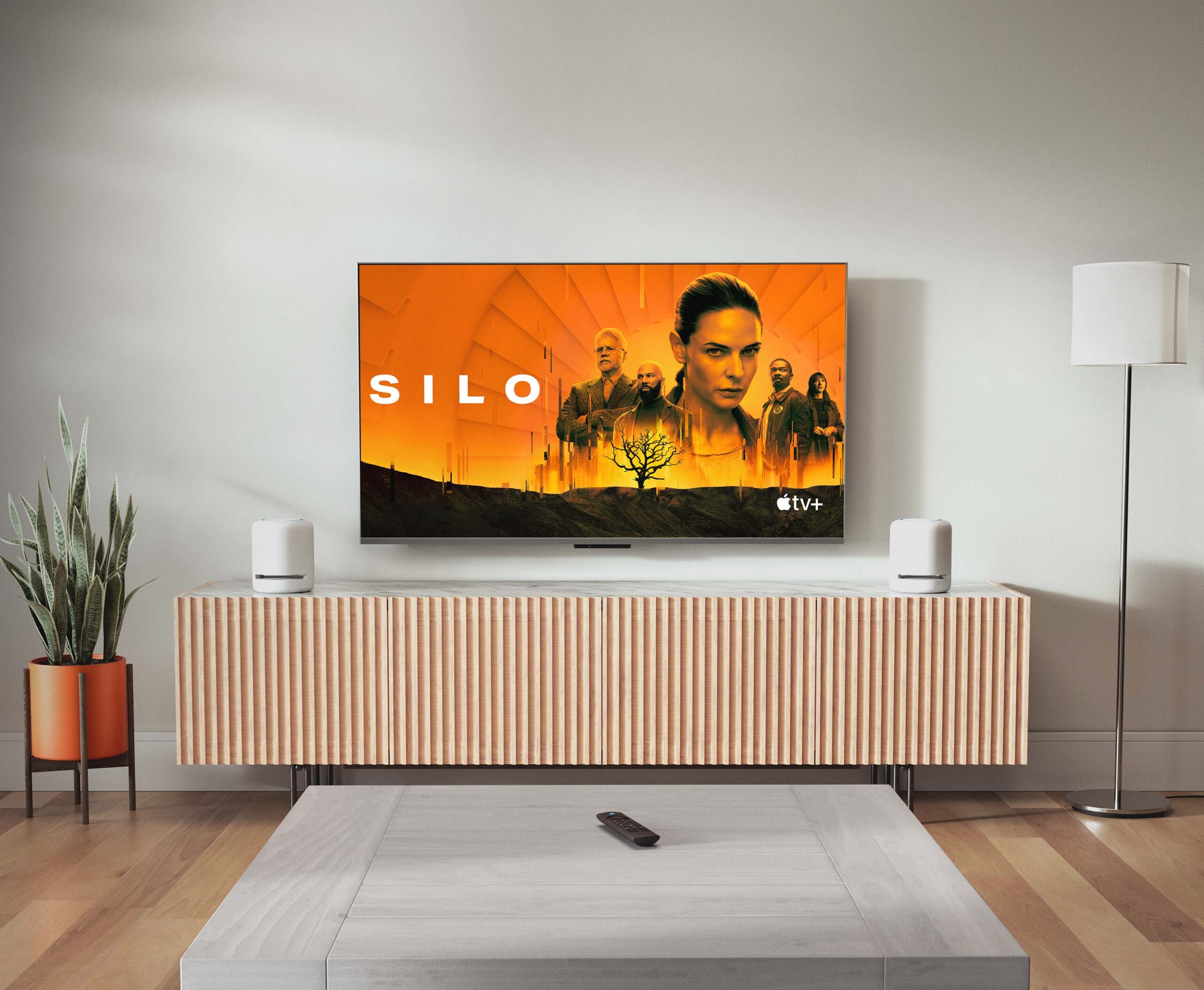 Una televisión de HD instalada en la pared de una sala muestra “Silo” de Apple TV+ junto a dos dispositivos Echo Studio: parlantes cilíndricos de 7 pulgadas de ancho y 8 pulgadas de alto y cuatro botones en relieve en la parte superior. 